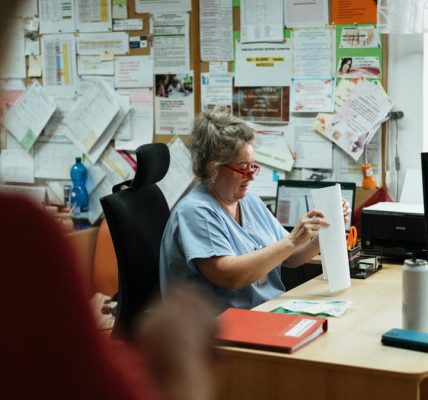 A senior caregiver in blue scrubs sorting paperwork at a hospital office desk.