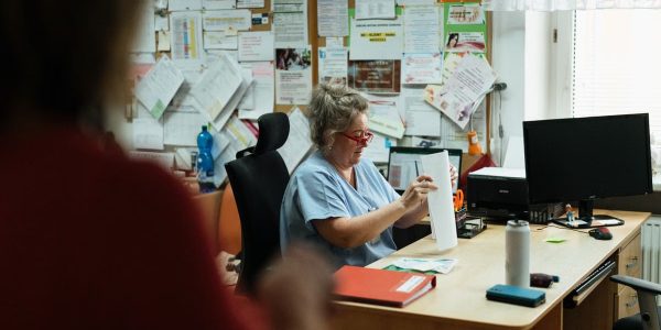 A senior caregiver in blue scrubs sorting paperwork at a hospital office desk.